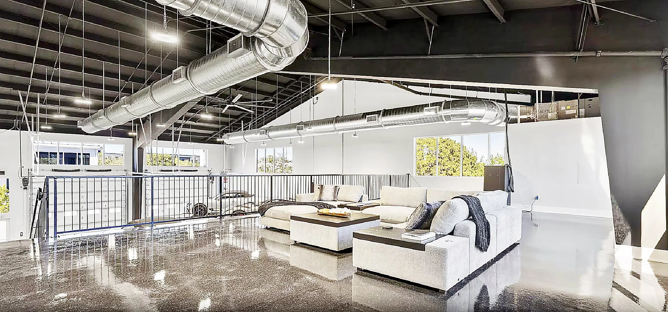 Modern loft apartment living room with exposed ductwork, white sectional sofa, coffee table, and large windows showing greenery outside.