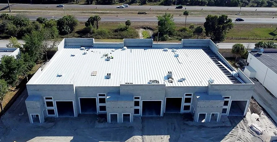 Aerial view of a flat rooftop building with multiple garage-style doors, under construction, with a highway and trees in the background.