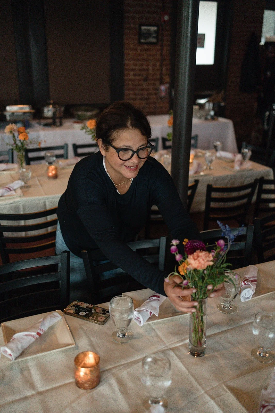 A woman with short dark hair and glasses arranging a flower bouquet on a dining table in a restaurant with brick walls and dim lighting.