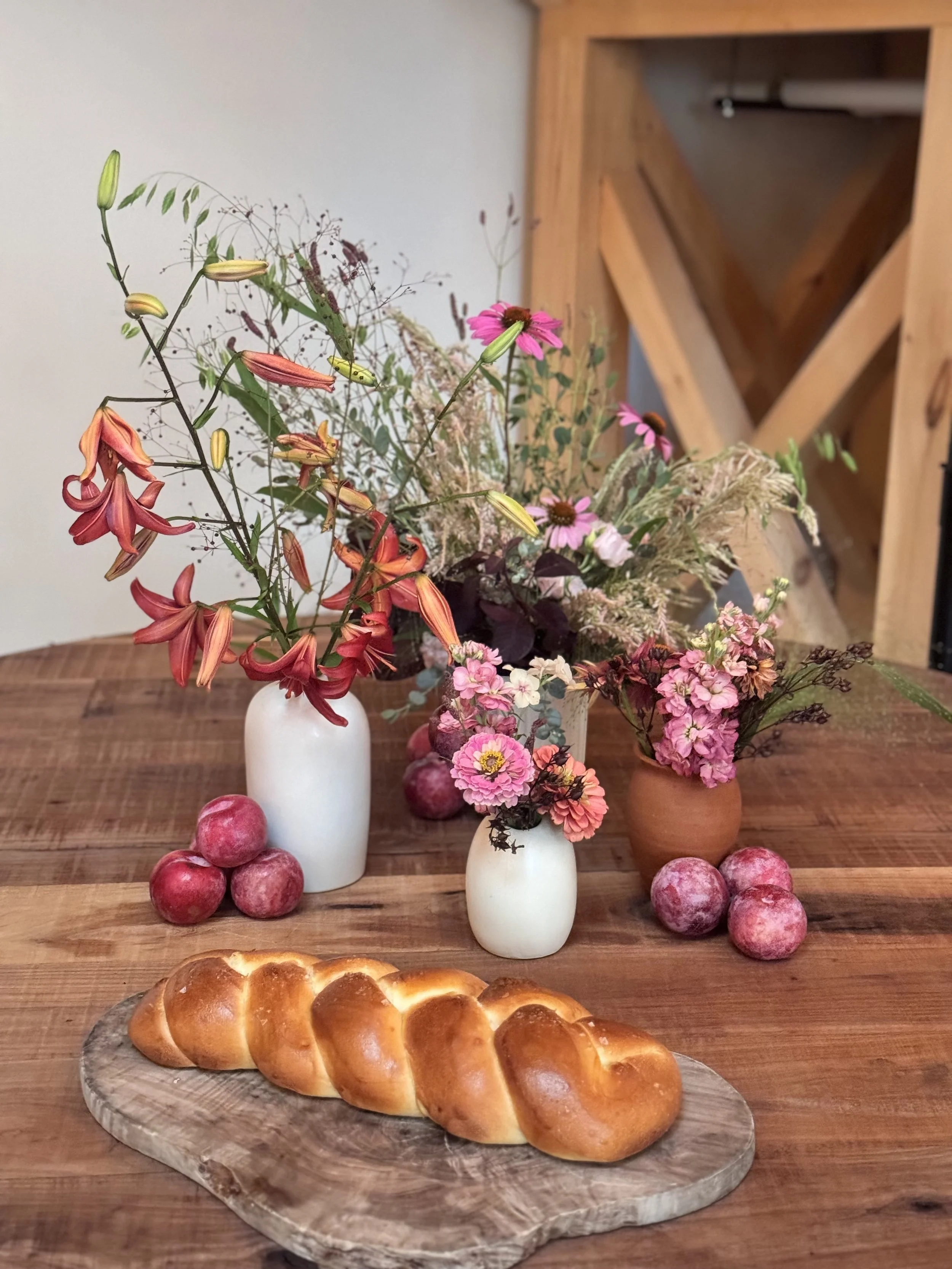 A wooden table with a loaf of bread on a wooden board, surrounded by three vases filled with various pink, red, and white flowers, and several red apples scattered on the table.