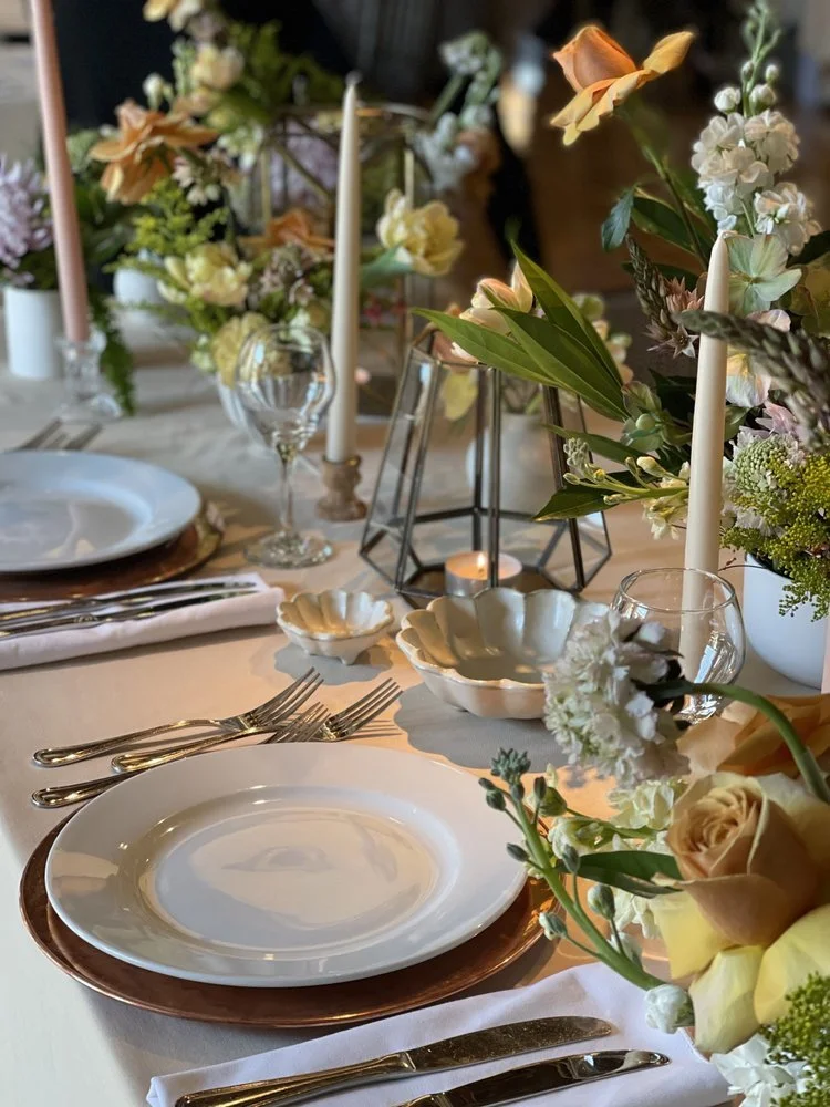 Elegant table setting with white plates on copper chargers, silverware, wine glasses, and floral centerpieces with pink, white, and green flowers, candles, and decorative bowls.