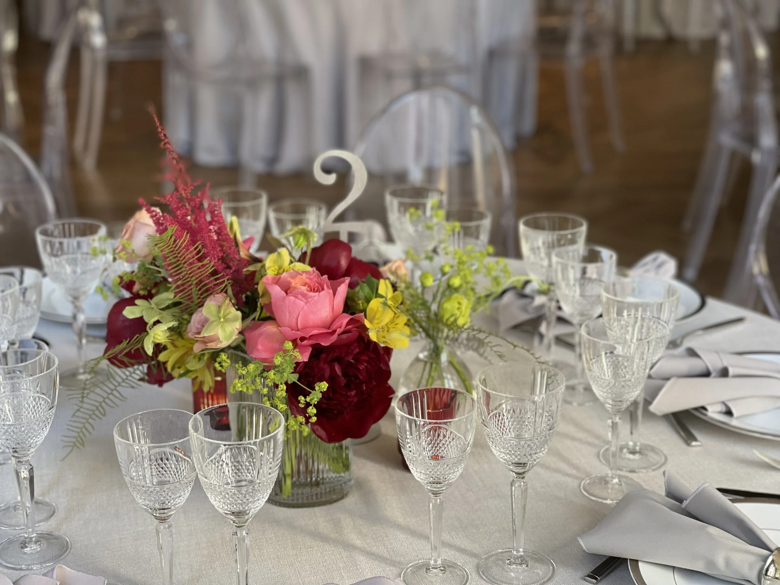 A round banquet table decorated with a floral centerpiece containing pink, red, and yellow flowers, surrounded by empty crystal glasses, white napkins, and place settings, in a formal event setting with additional empty chairs in the background.