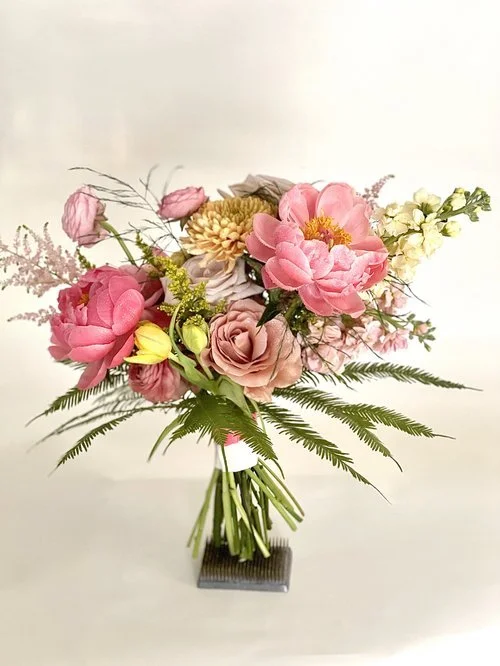 A bouquet of pink, yellow, and beige flowers with green foliage in a clear glass vase on a small black square stand.