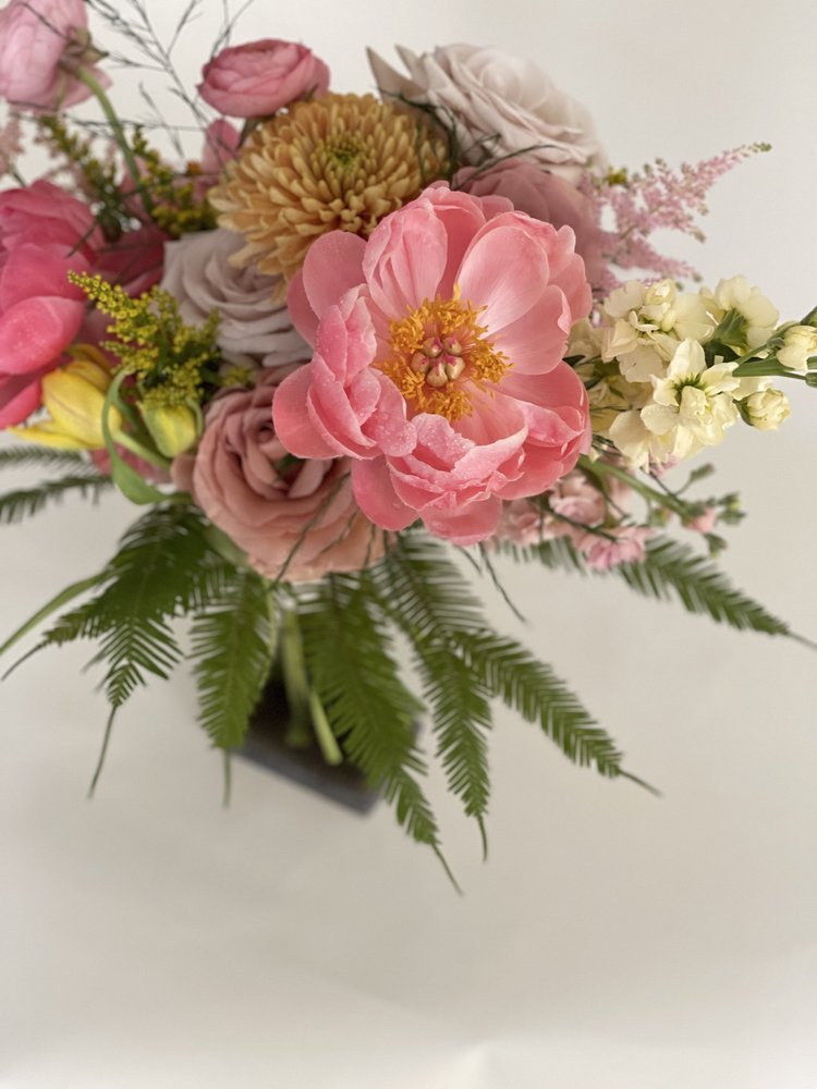 A colorful bouquet of various flowers including pink, yellow, purple, and white blossoms, arranged with green fern leaves in a black square vase.