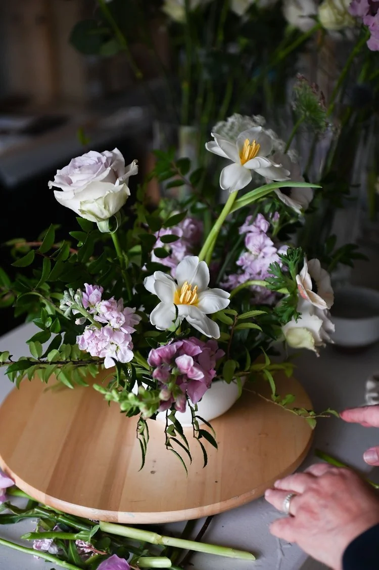 Person arranging a bouquet of white, pink, and purple flowers in a white vase on a wooden tray.
