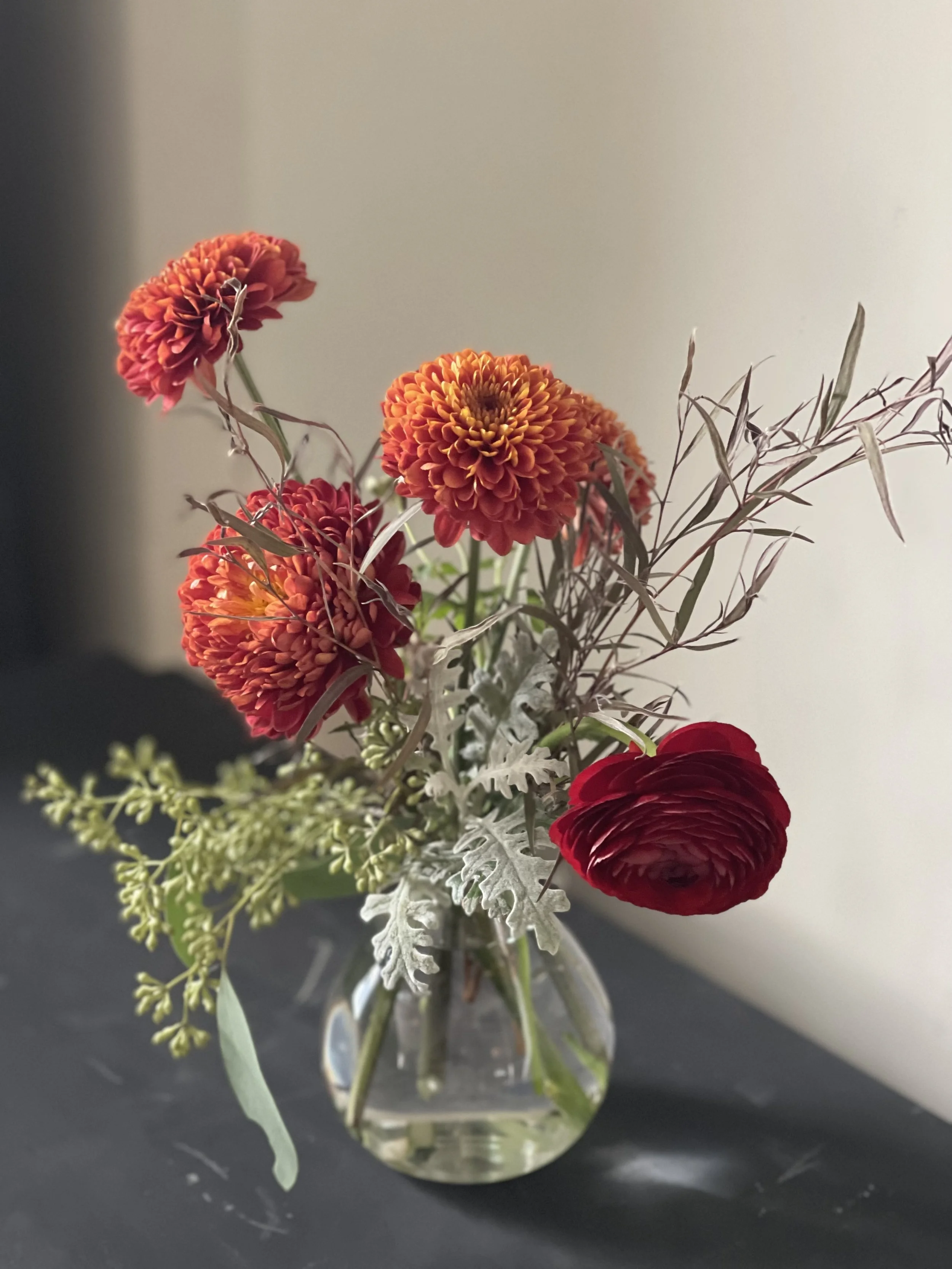 A flower arrangement in a glass vase with orange, red, and burgundy flowers along with green and grayish foliage, placed on a black surface against a light background.