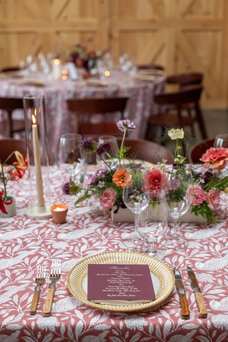 A decorated dining table with a pink floral tablecloth, floral centerpiece, candle, silverware, and wine glasses in a rustic wooden room.