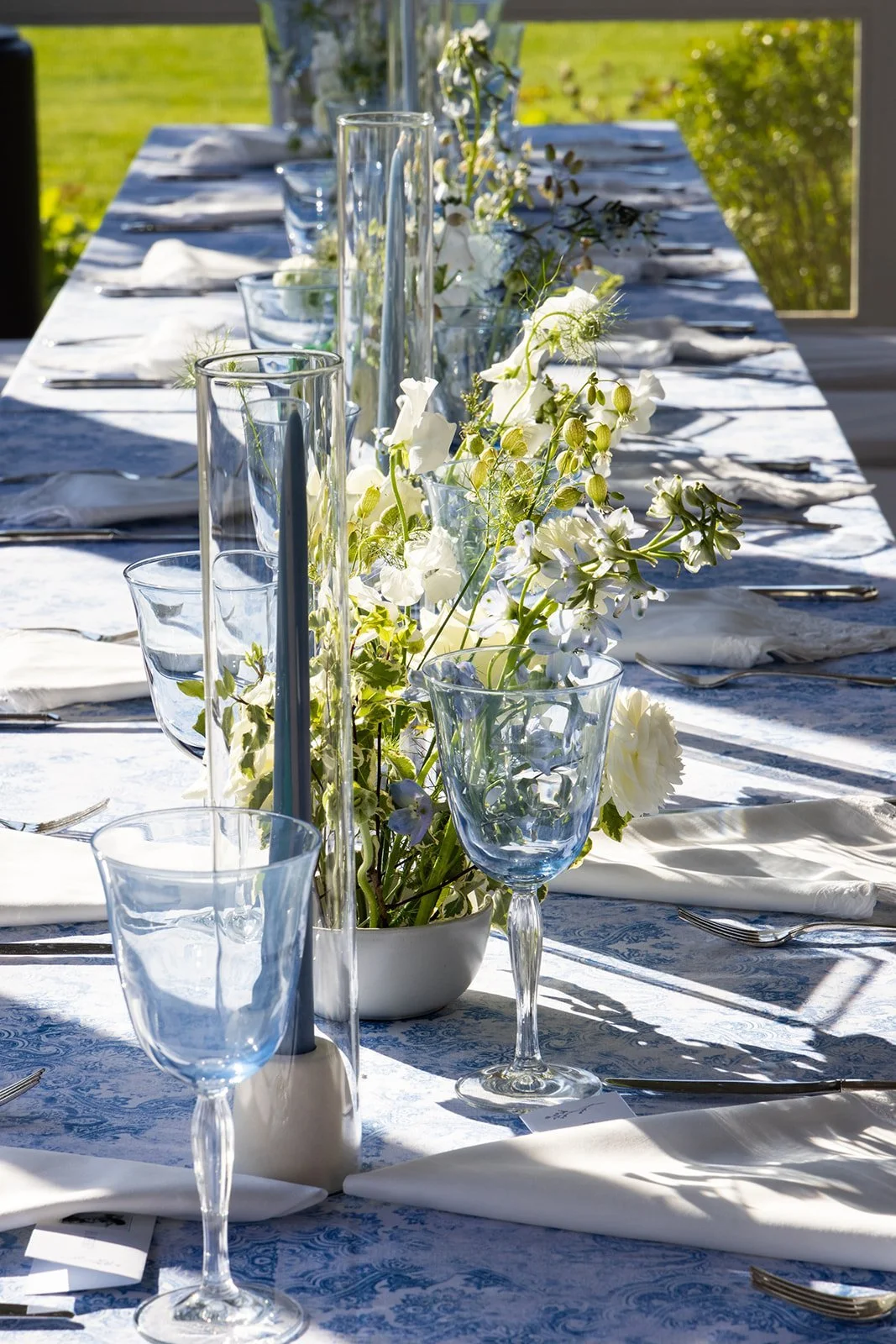 A long outdoor dining table decorated with white and blue tablecloth, white napkins, glassware, silverware, and a floral centerpiece in a white vase on a sunny day.