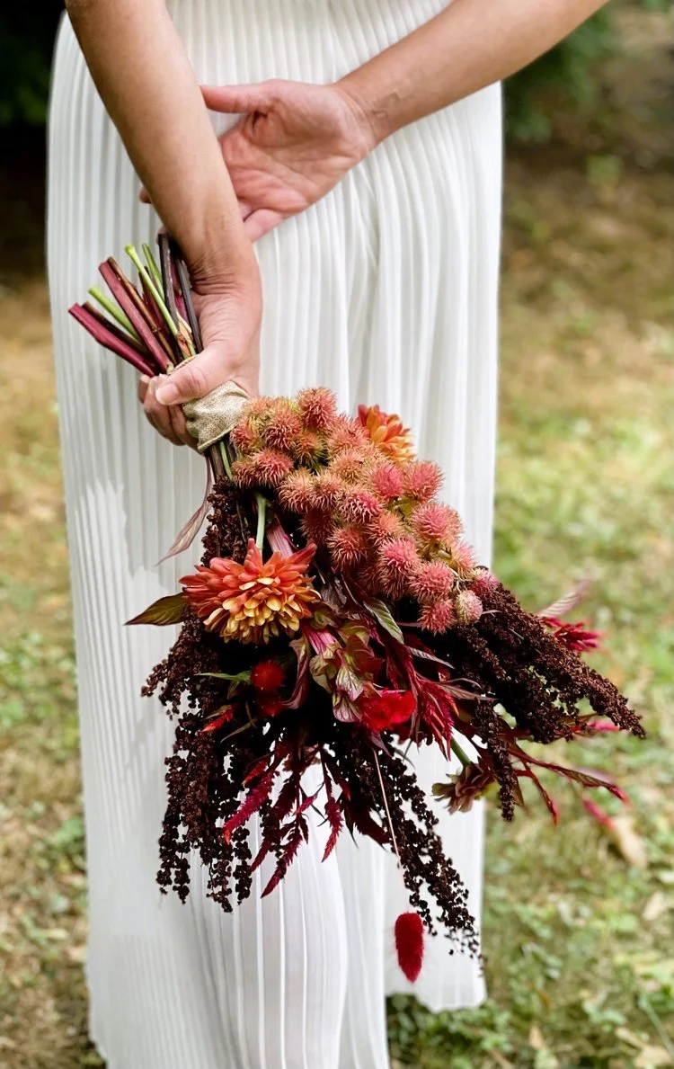 A person in a white pleated dress holding a colorful flower bouquet, outdoors with grass and blurred greenery in the background.
