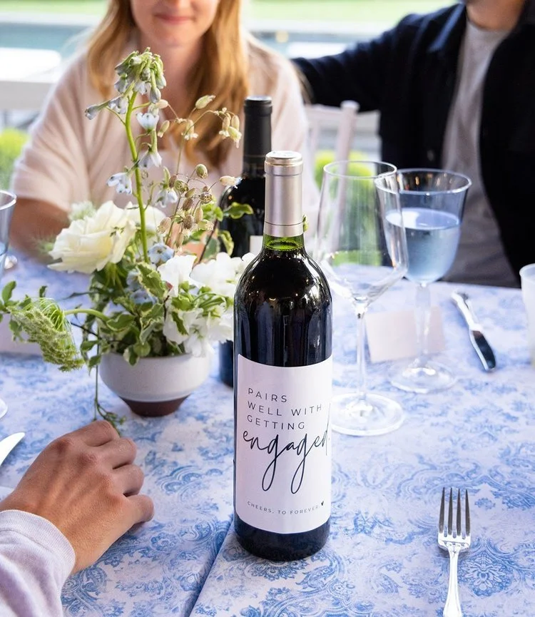 A wine bottle with a decorated label that says 'Pairs well with getting engaged', surrounded by wine glasses, a floral centerpiece, and people at a table during a celebration.