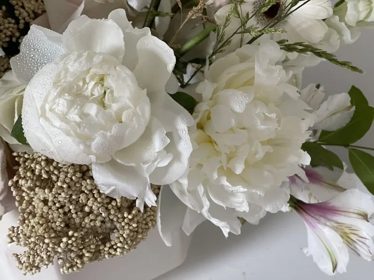 Close-up of white peonies and other white flowers with green leaves, some with water droplets, arranged in a bouquet.