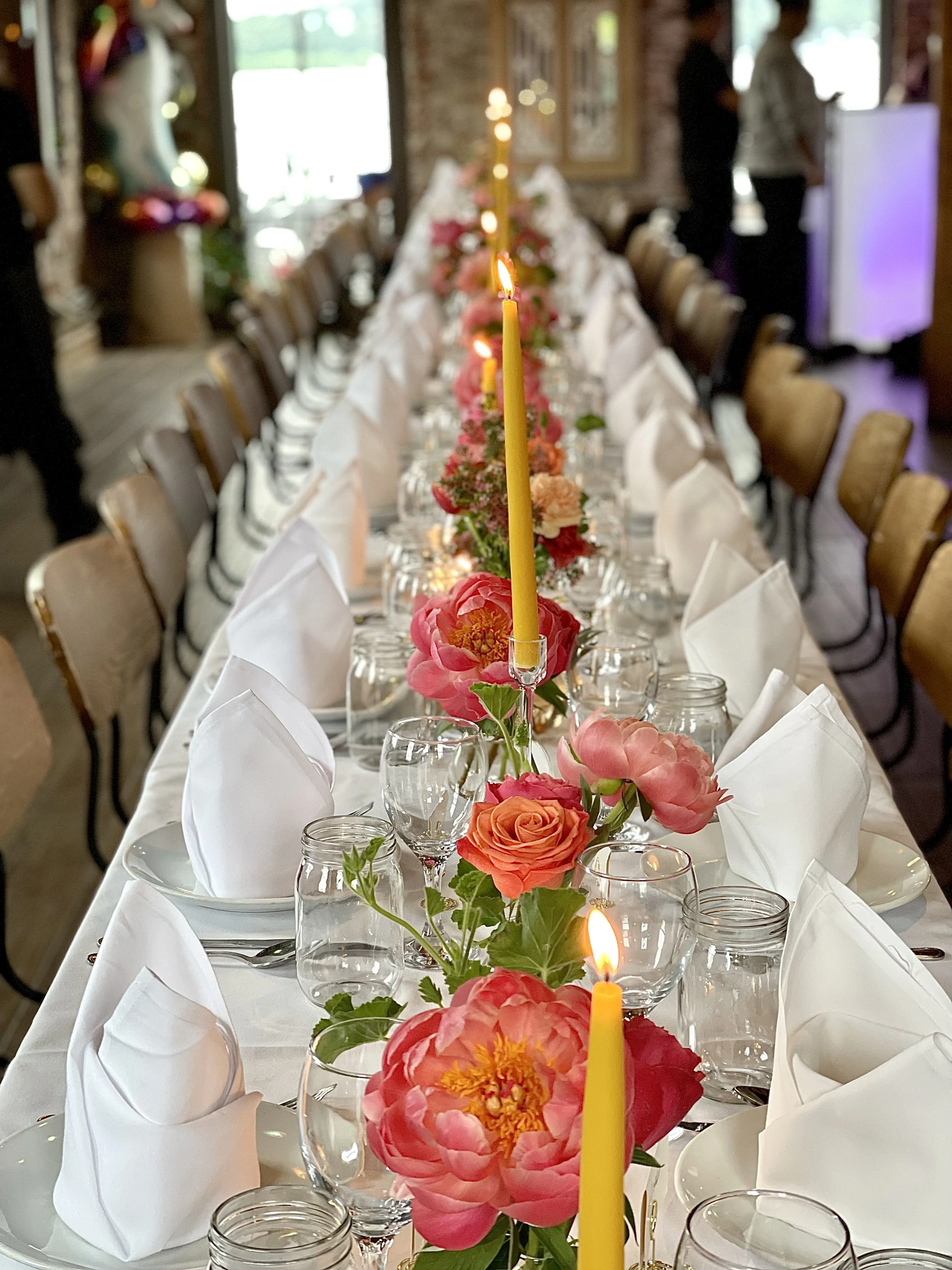 A long banquet table decorated with pink and orange flowers, white napkins, glassware, candles, and brown chairs in a cozy indoor setting for a special event.