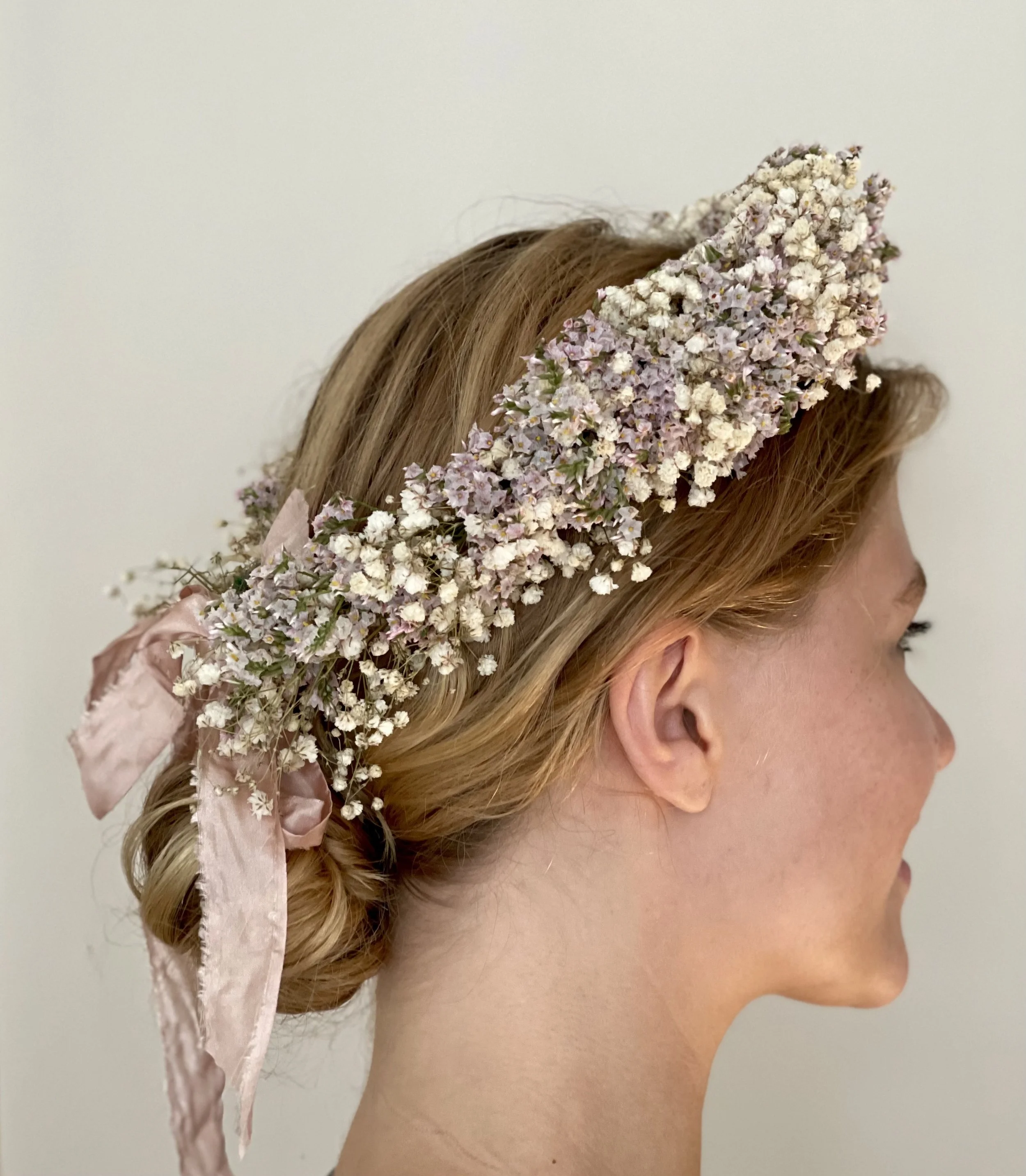 A woman with red hair wearing a floral crown made of small white and pastel purple flowers, tied with a pink satin ribbon at the bun.