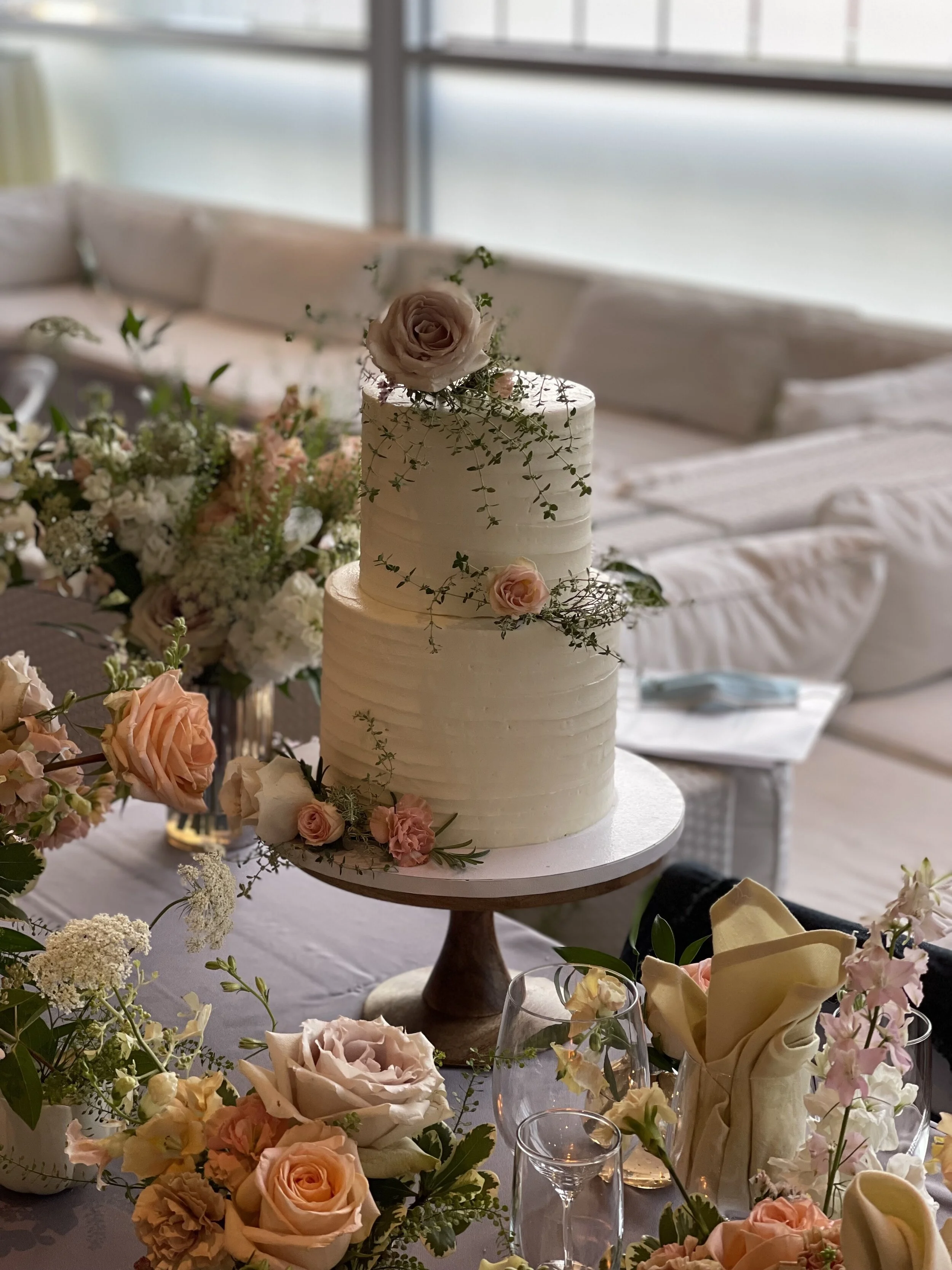 A two-tiered white wedding cake decorated with pink and cream roses and greenery, placed on a wooden cake stand at a wedding reception with floral arrangements, glasses, and napkins on the table.