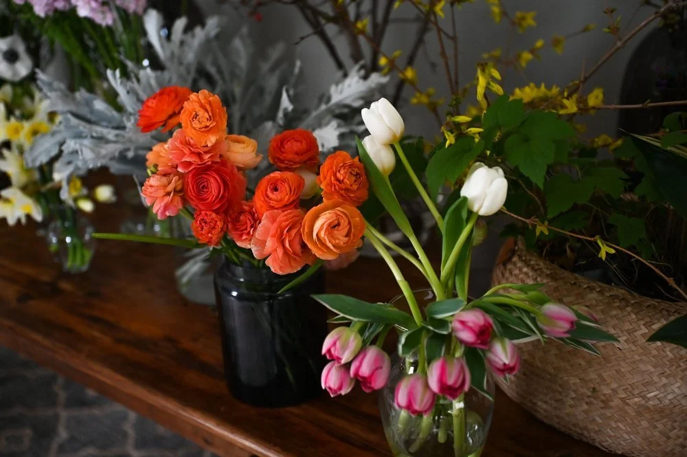 Vases of colorful flowers on a wooden table, including orange and pink ranunculus, white tulips, and pink-tipped tulips, with other foliage in the background.