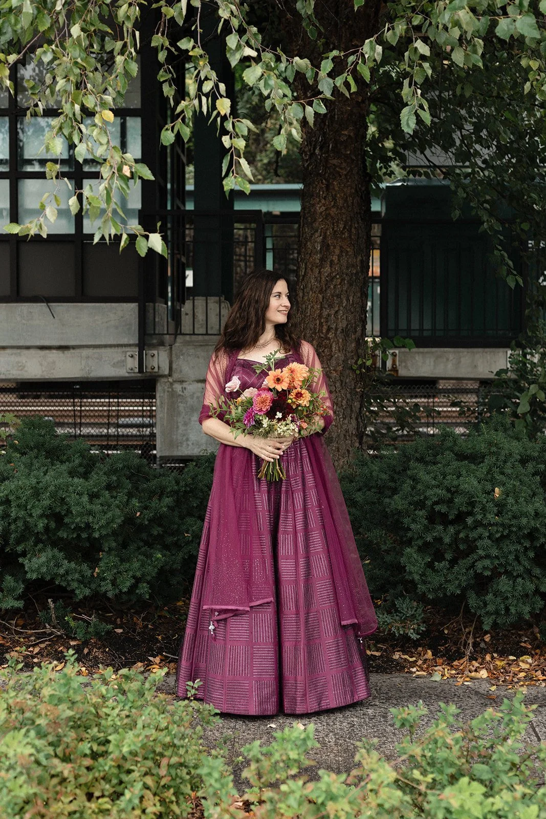 A woman in a long purple dress holding a bouquet of flowers, standing outdoors next to a tree with green foliage.