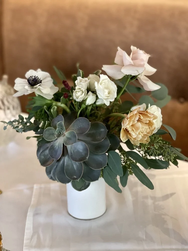A floral arrangement in a white vase featuring white roses, peach peony, anemone, succulents, and green foliage on a white cloth table.