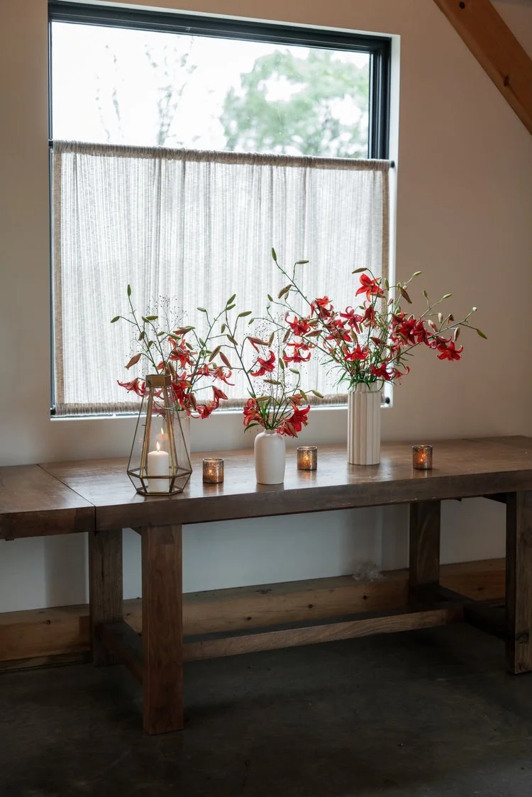 A wooden table with three vases and four candles in glass holders, positioned beneath a window with a beige curtain and scenic outdoor view.