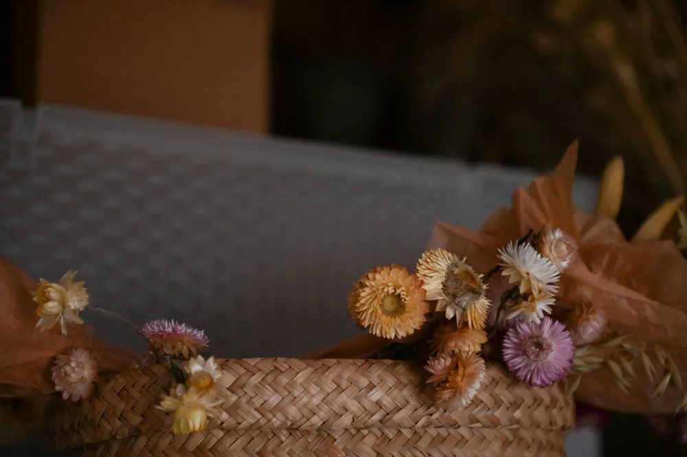 A close-up of dried flowers in shades of yellow, orange, and pink arranged in a woven basket with brown paper wrapping.