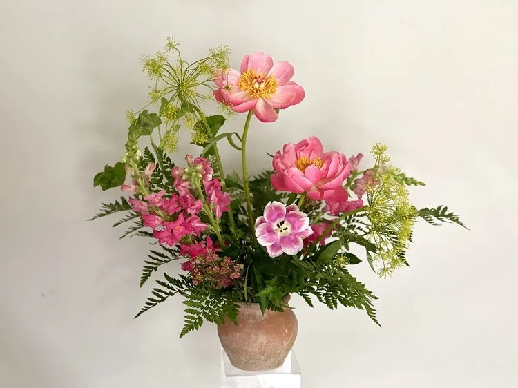 A bouquet of pink flowers, including peonies and primroses, with greenery in a pinkish vase on a white surface against a plain white background.