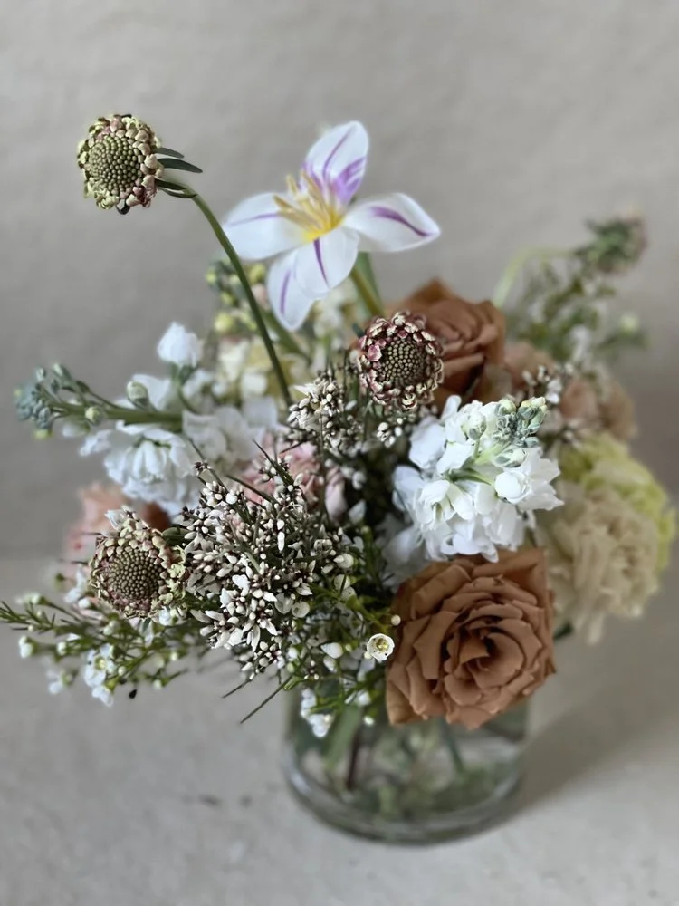 A bouquet of mixed flowers in a clear glass vase, including white, brown, and pink blooms.