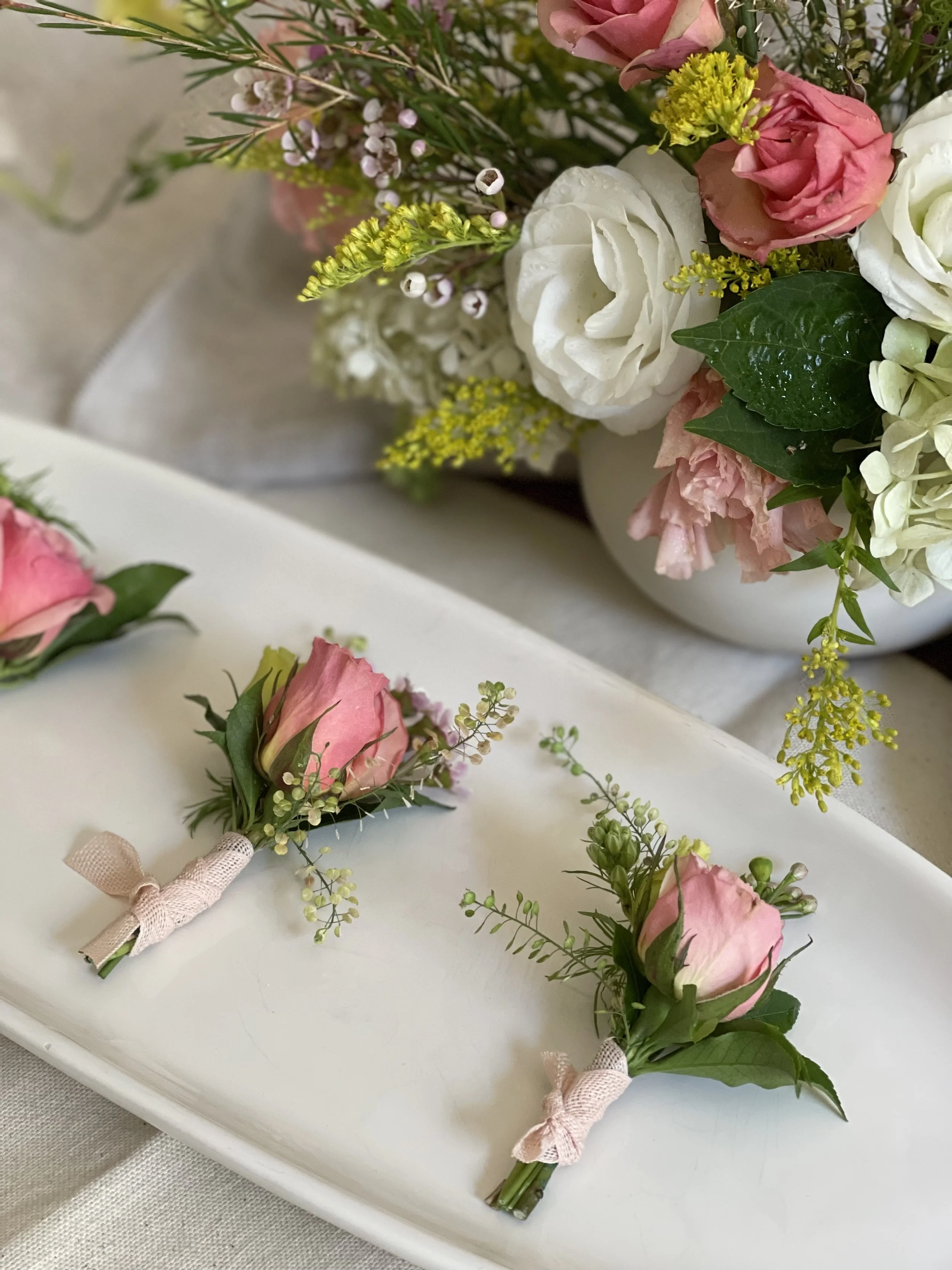 Close-up of a floral arrangement featuring white, pink, and yellow flowers with green leaves, and two small boutonnières with pink roses and greenery on a white tray.