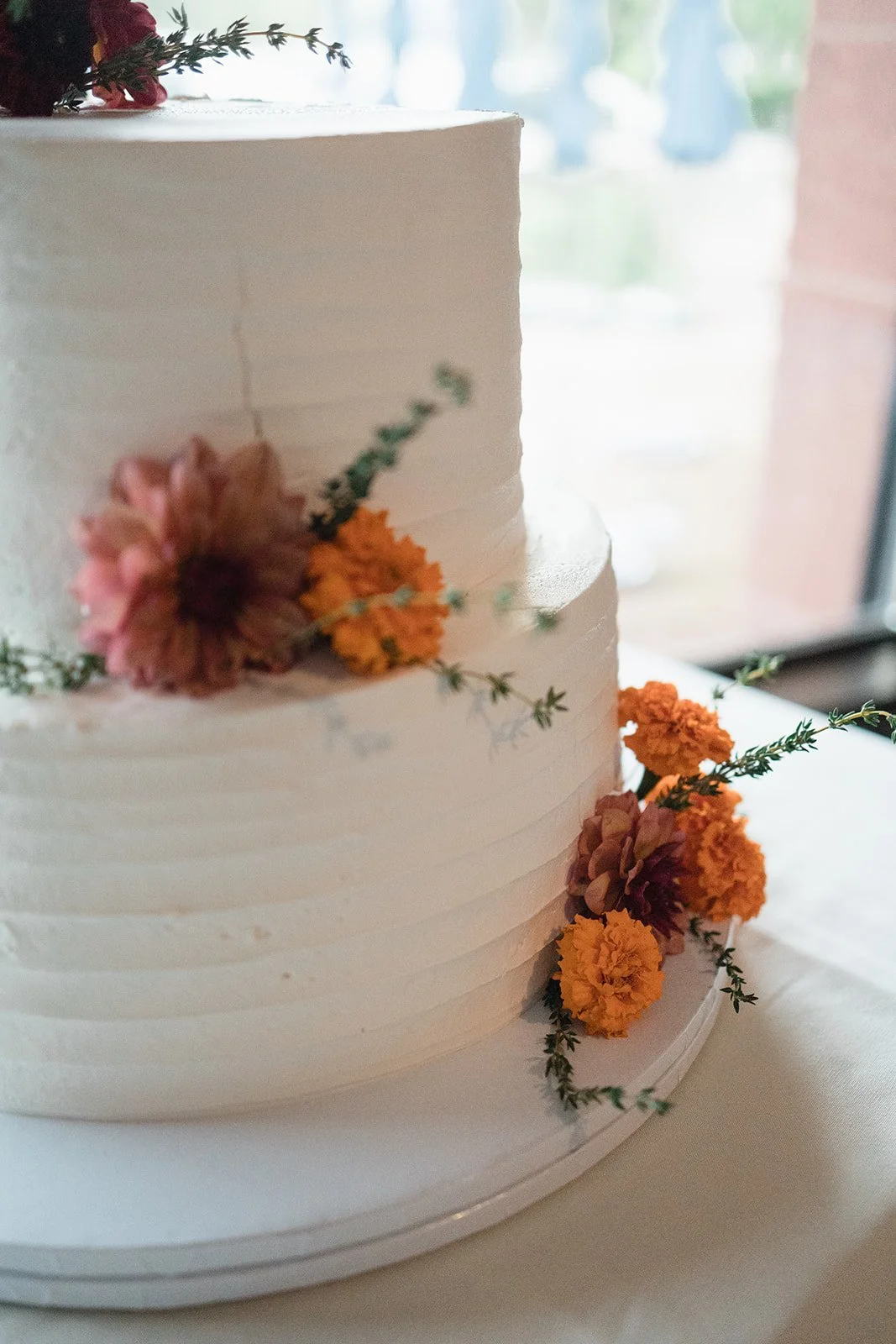 A white, multi-layered wedding cake decorated with pink and orange flowers and green sprigs, placed on a white cake stand near a window.