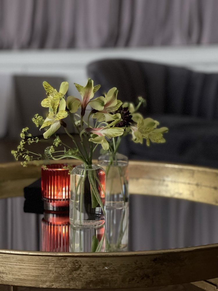A floral arrangement in three small vases on a round table with a mirror surface, with a red candle holder and a candle inside. In the background, a dark sofa and curtains are visible.