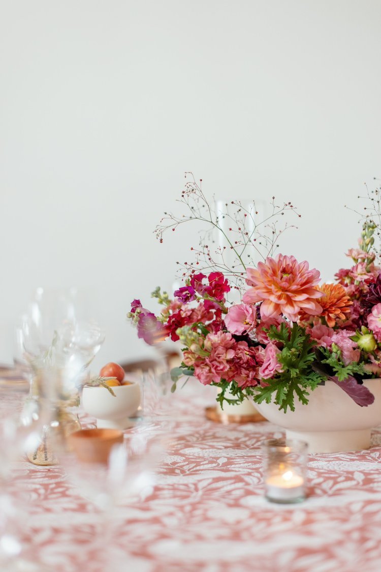 A floral centerpiece with pink, orange, and purple flowers on a table with a pink floral tablecloth, surrounded by small candle holders and bowls.