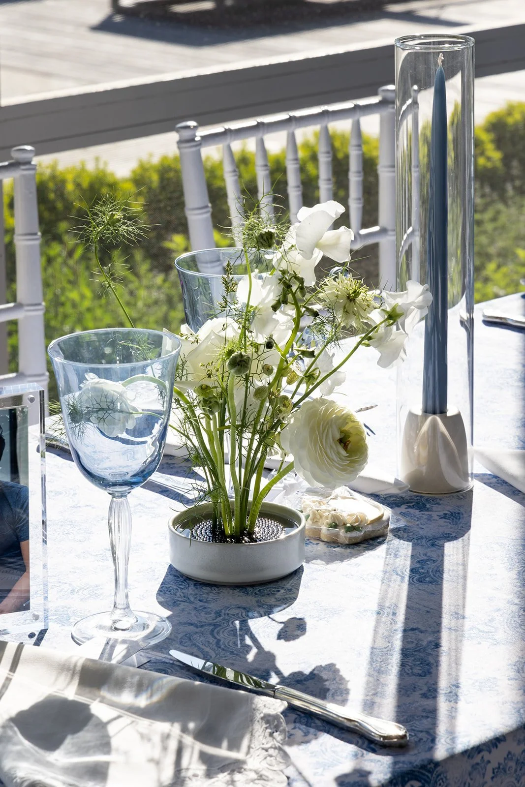 A decorated outdoor table with a white tablecloth, a small white flower arrangement, tall blue candle in a glass holder, two clear glass vases with water, and silverware. Sunlight casts shadows on the table.