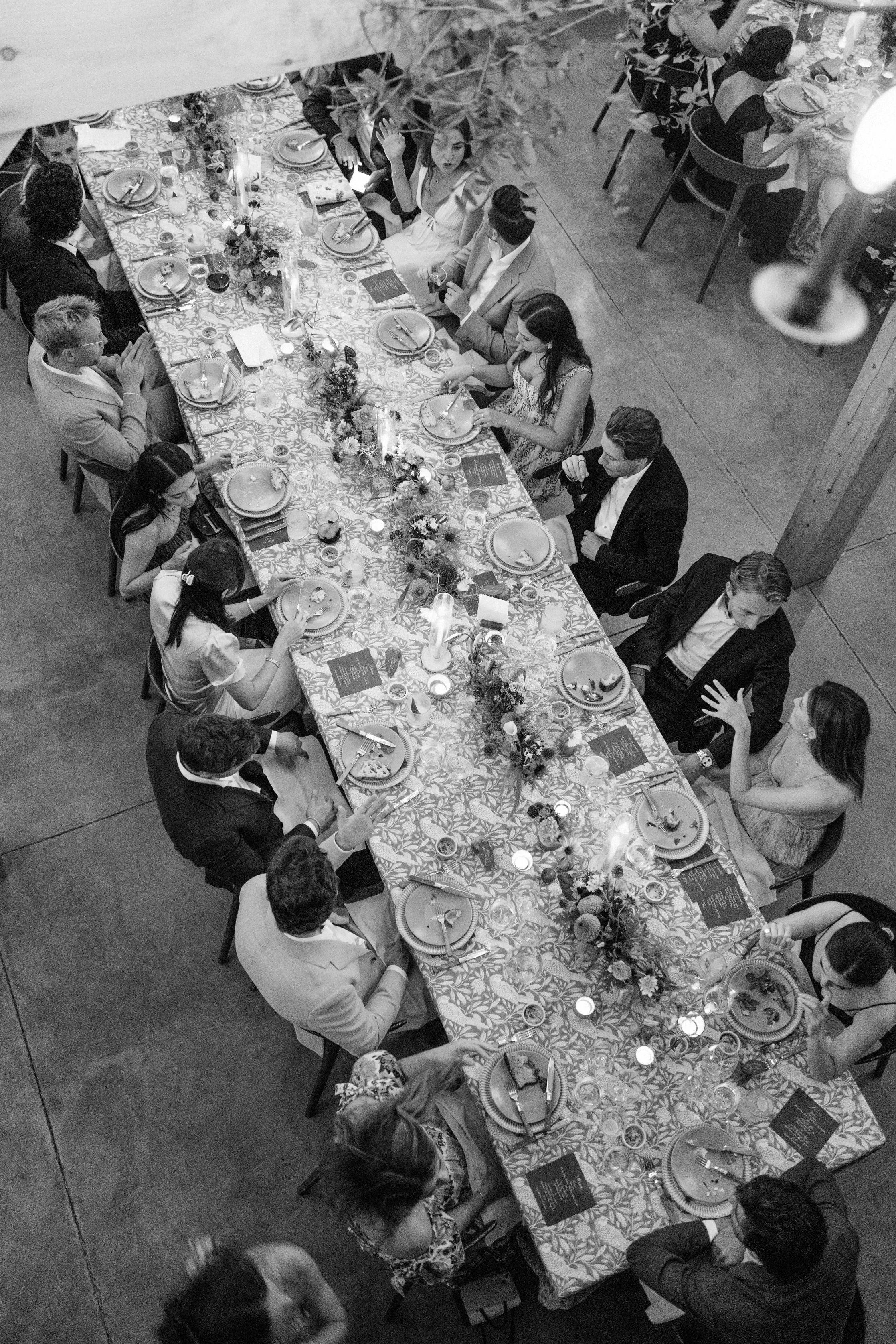 A black-and-white photo of a banquet with people seated around a long decorated table set with plates, glasses, and centerpieces.