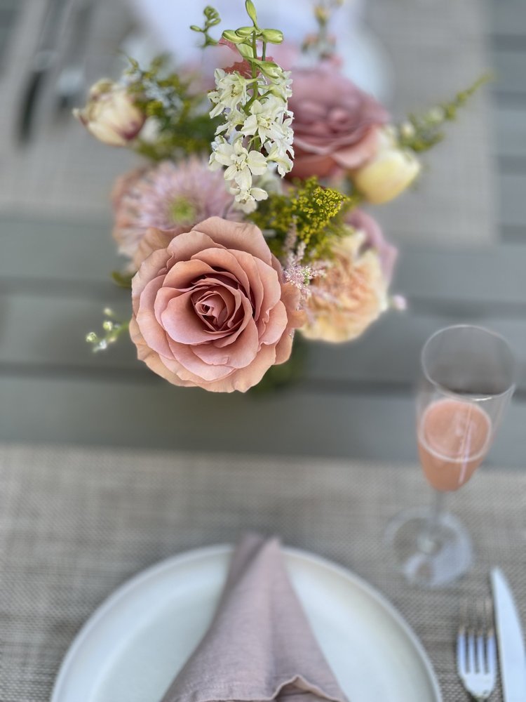 Close-up of a floral centerpiece with pink, white, and cream roses, and other flowers on a dining table set with a plate, napkin, fork, knife, and glass of pink drink.