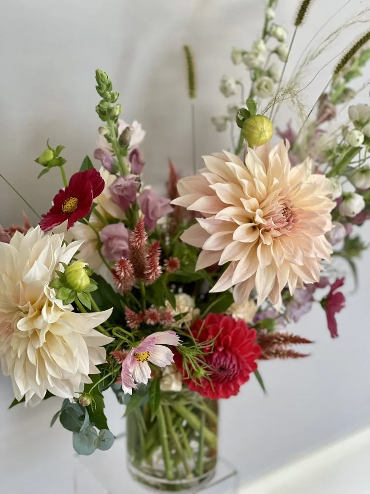 A colorful bouquet of assorted flowers in a clear glass vase, including dahlias, snapdragons, and other blossoms, on a white surface.