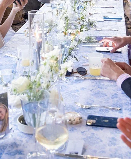 A formal dining table set for a celebration with white flowers, glasses of wine, and candles, with people using their phones.