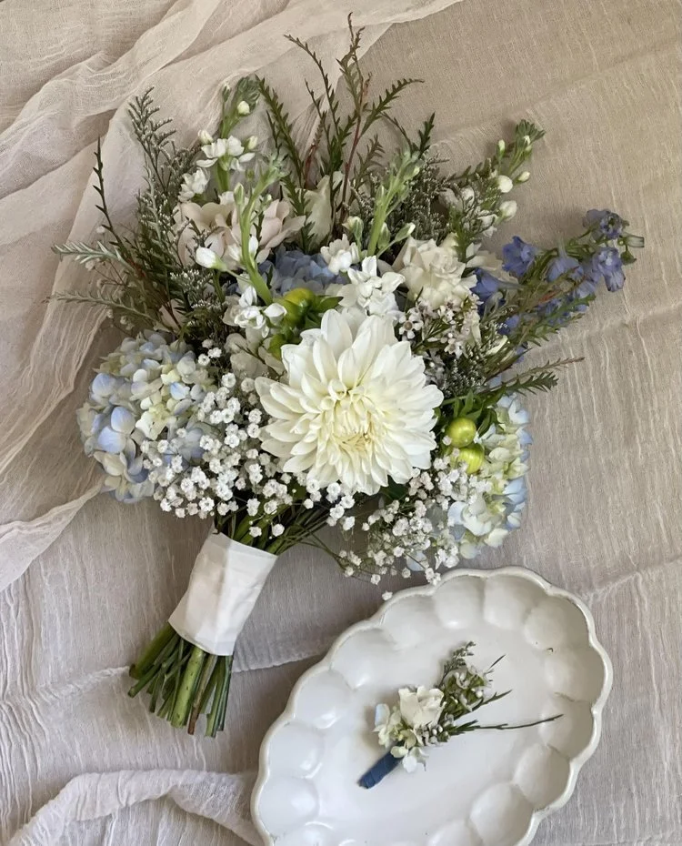 A bouquet of mixed white, blue, and green flowers with greenery, tied with a white ribbon, resting on a light-colored fabric. There is also a white ceramic plate holding a small boutonniere with matching flowers and greenery.