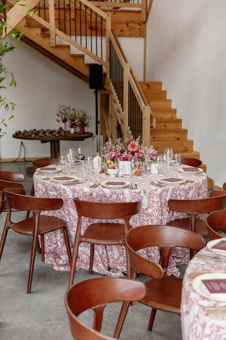 Round dining table with a pink floral tablecloth set with plates, glassware, napkins, and a floral centerpiece. Several wooden chairs are around the table. In the background, there is a wooden staircase and a table with flowers and fruits on it.