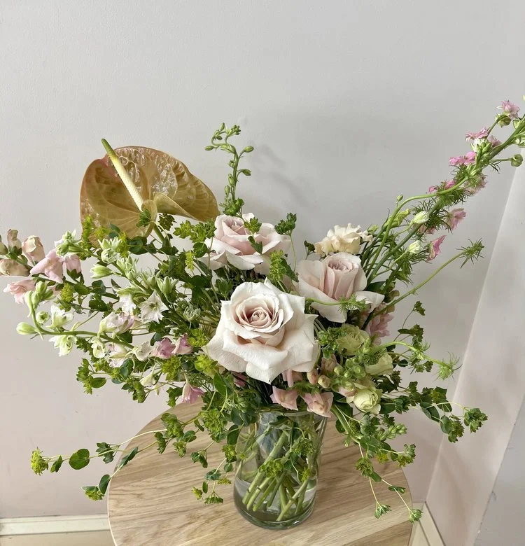 A glass vase with a bouquet of white and pink roses, green foliage, and various wildflowers, placed on a wooden surface against a plain white wall.