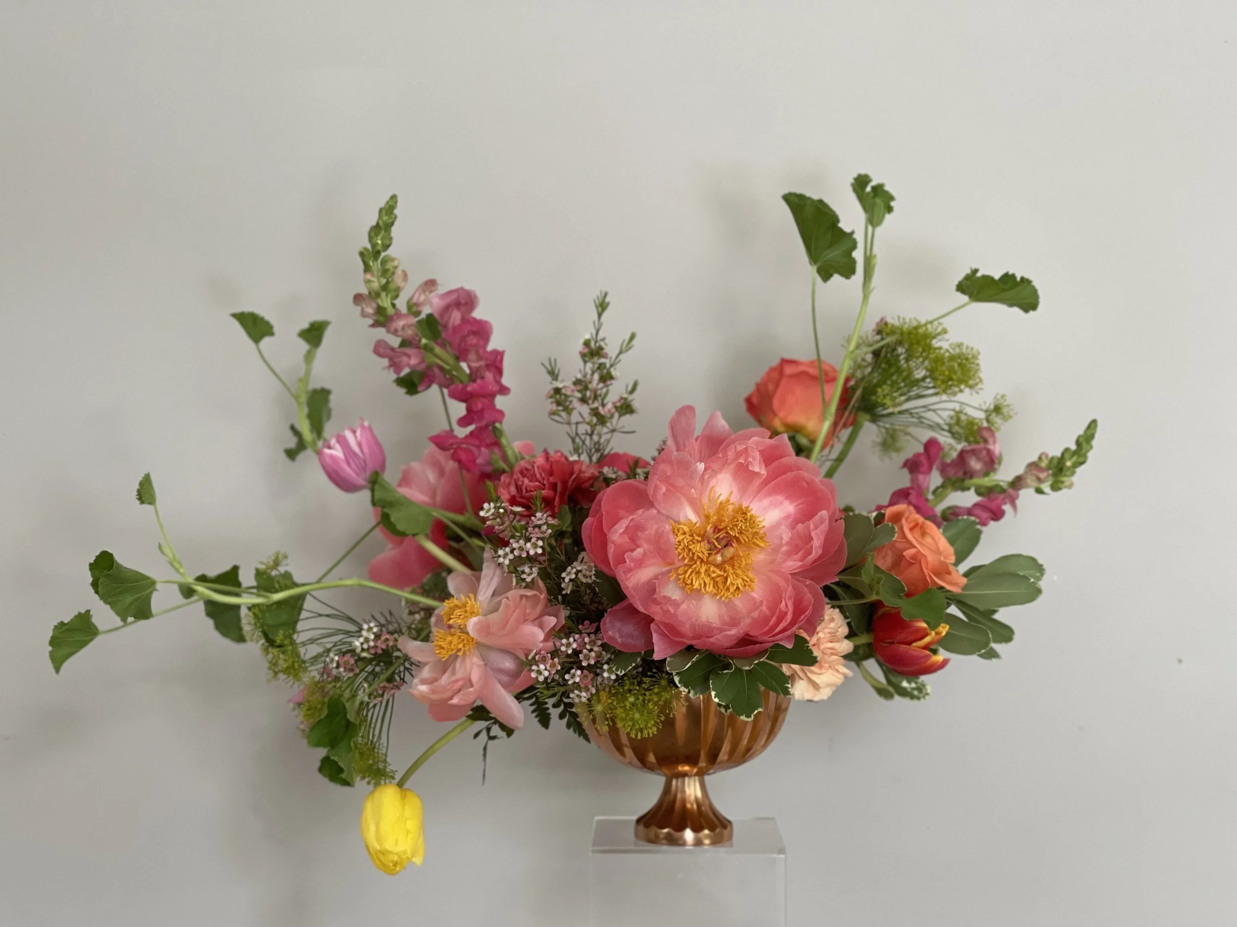 Colorful floral arrangement in a copper vase with pink, orange, and yellow flowers and green foliage on a plain white background.