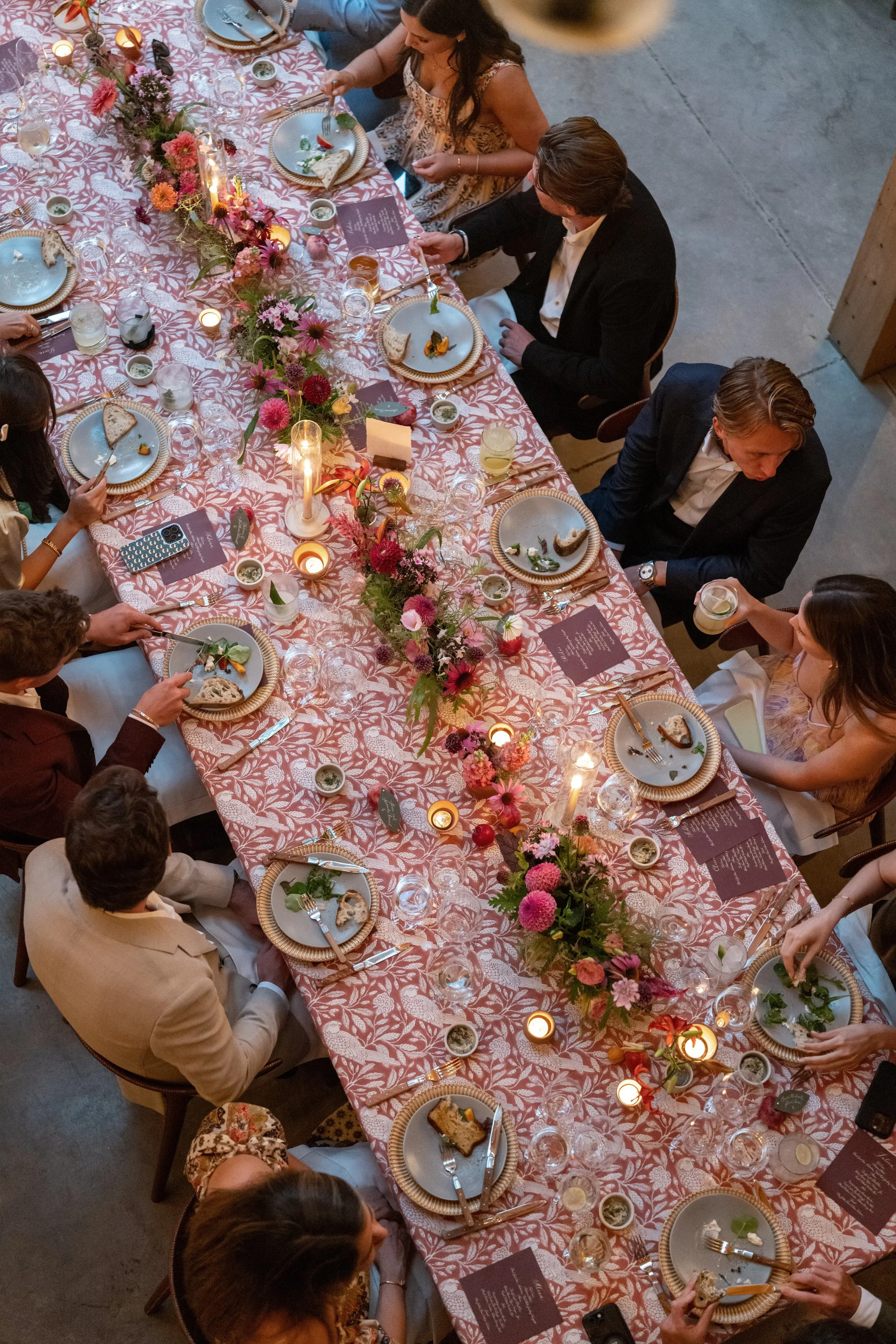 A top-down view of a large, festive dinner table set with plates, utensils, glasses, and a vibrant floral centerpiece. Several guests are seated, some eating, some engaged in conversation, and one person serving drinks. The table is decorated with ca