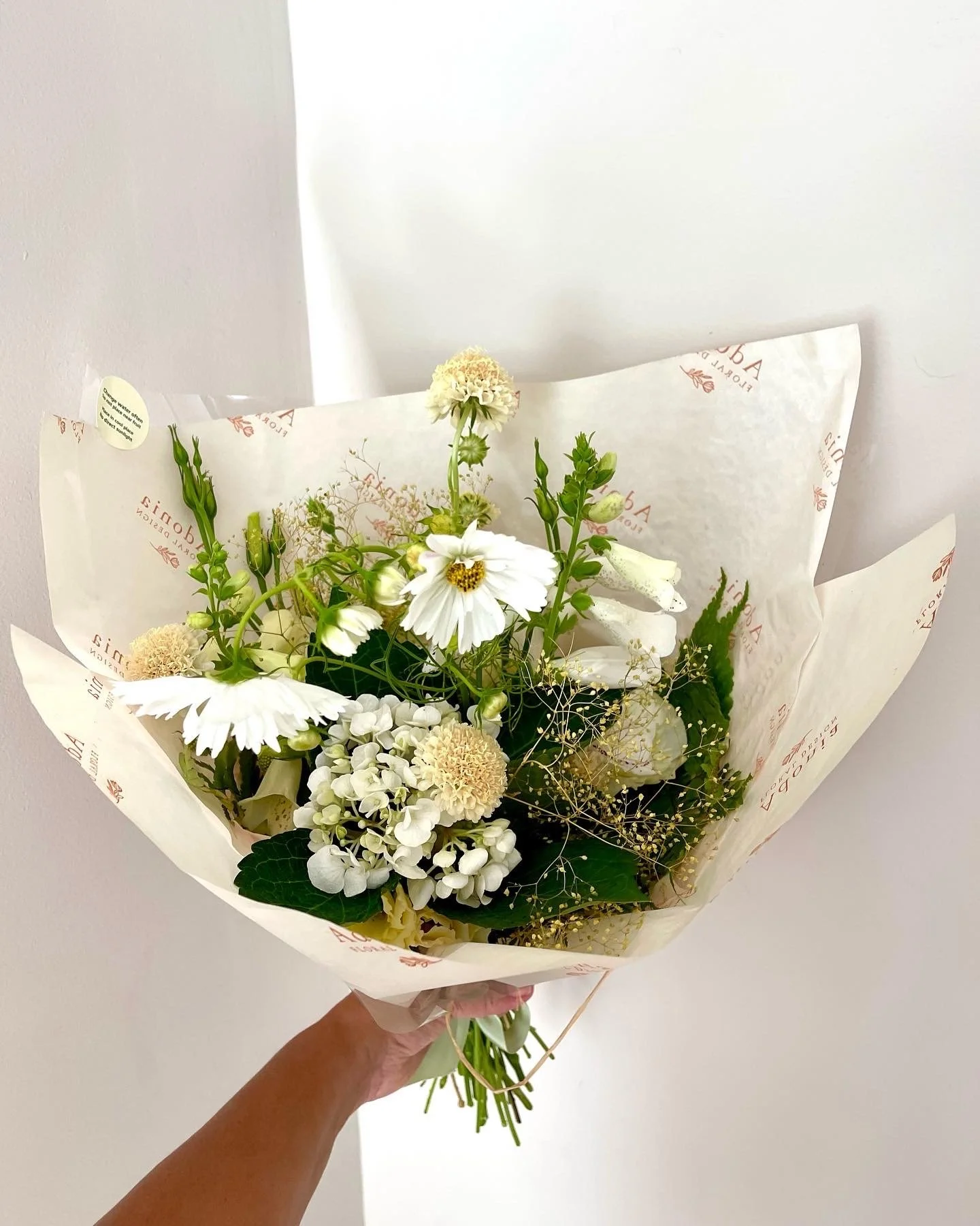 A bouquet of white and cream flowers arranged with greenery, wrapped in white paper with red text.