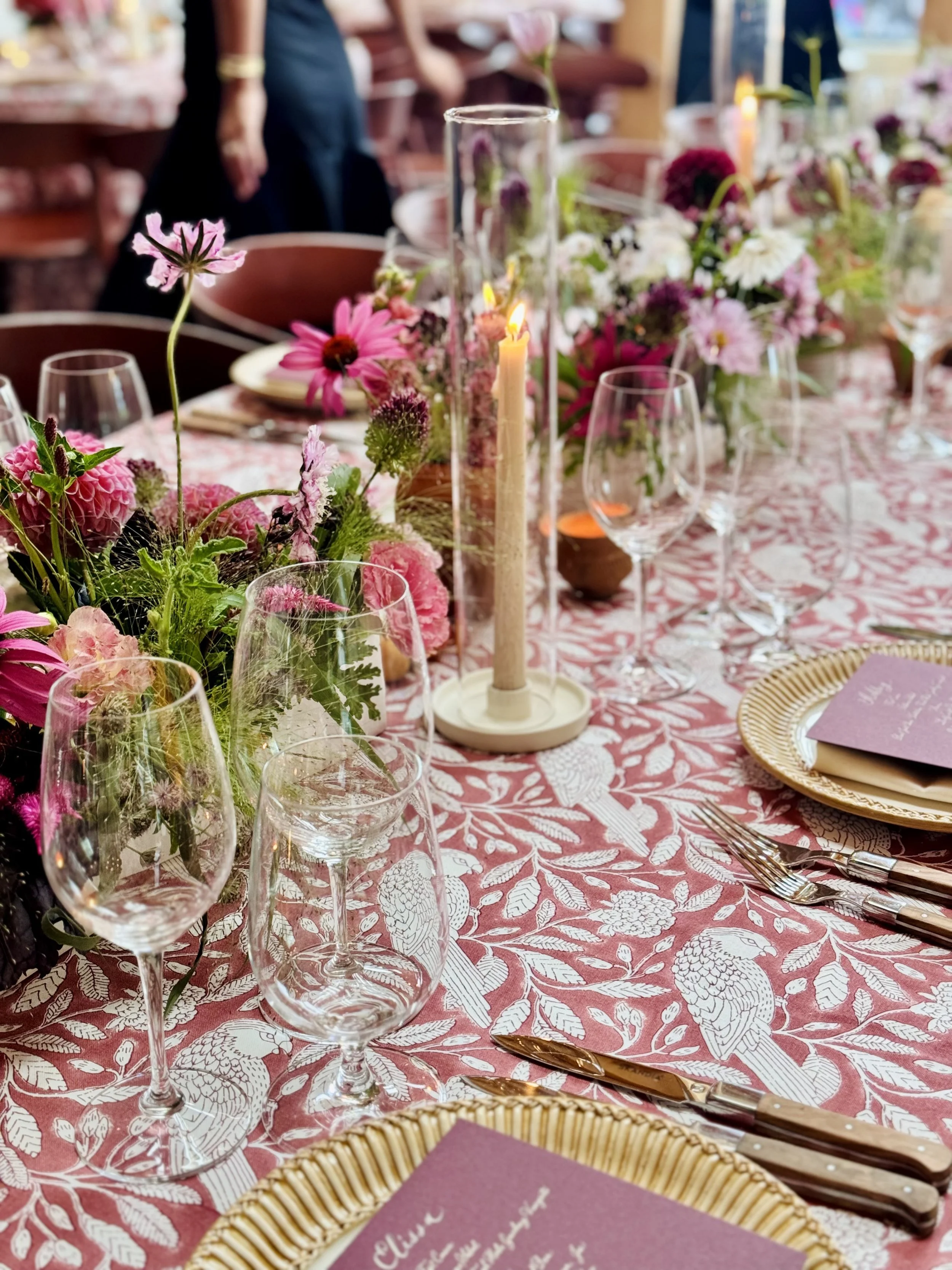 A table decorated for a celebration with pink and purple flowers, a lit candle in a glass holder, wine glasses, gold-rimmed plates, silverware, and purple napkins with writing, on a pink patterned tablecloth.