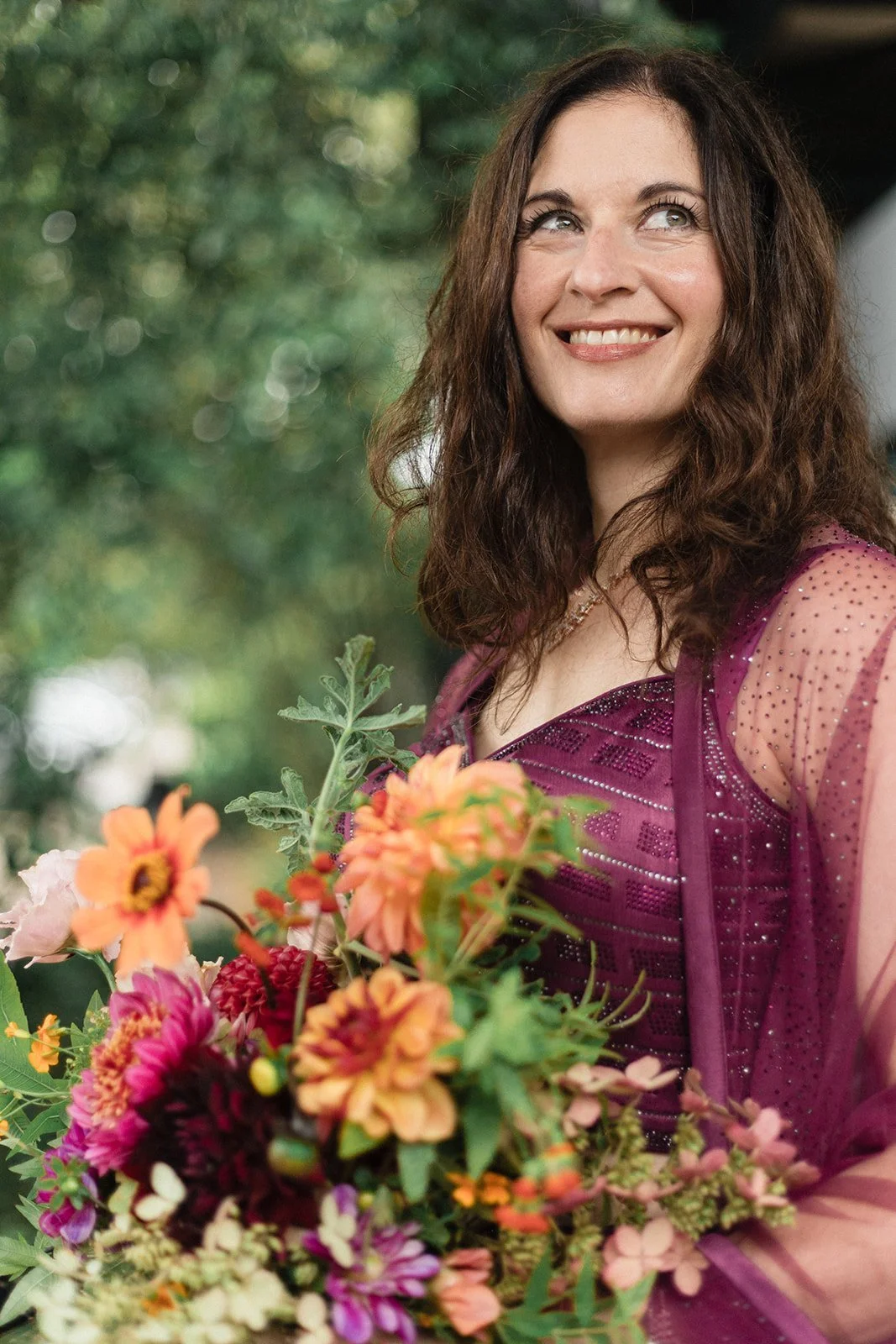 A woman with long curly brown hair smiling and looking away, holding a bouquet of colorful flowers outdoors with blurred green foliage in the background.