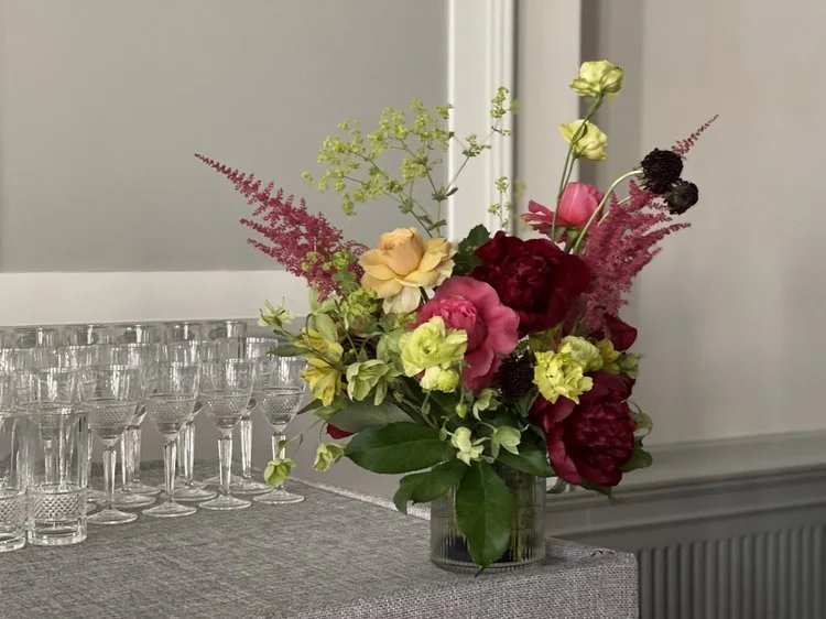 Colorful floral arrangement in a glass vase with pink, yellow, and burgundy flowers on a table with empty wine glasses nearby.
