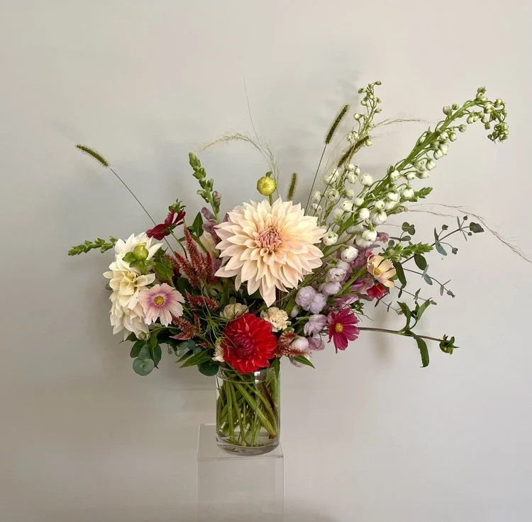 A colorful bouquet of mixed flowers in a glass vase on a clear stand against a plain wall.