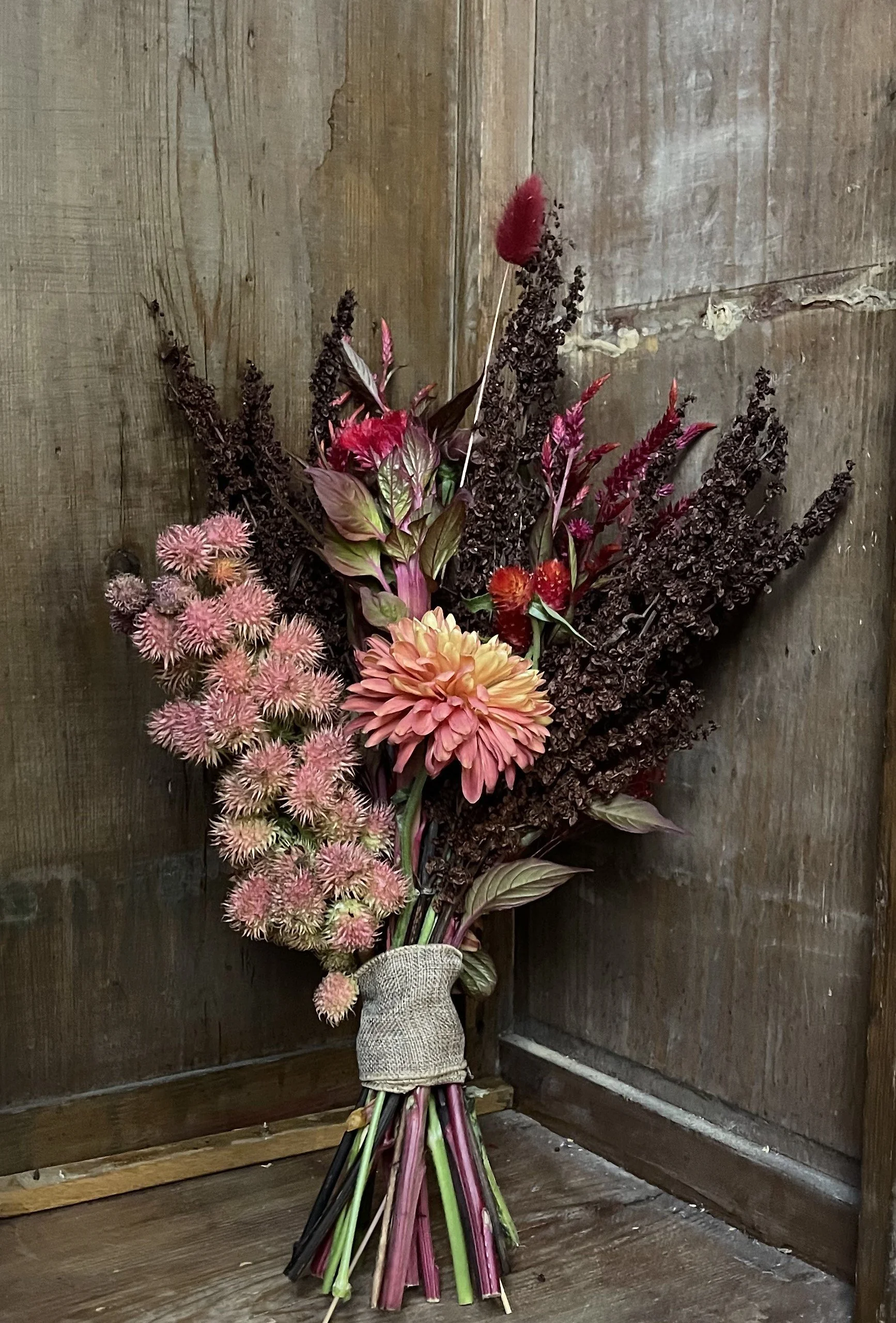 A bouquet of mixed flowers including pink blush, peach, and dark red blooms, wrapped with a burlap ribbon, leaning against wooden wall.