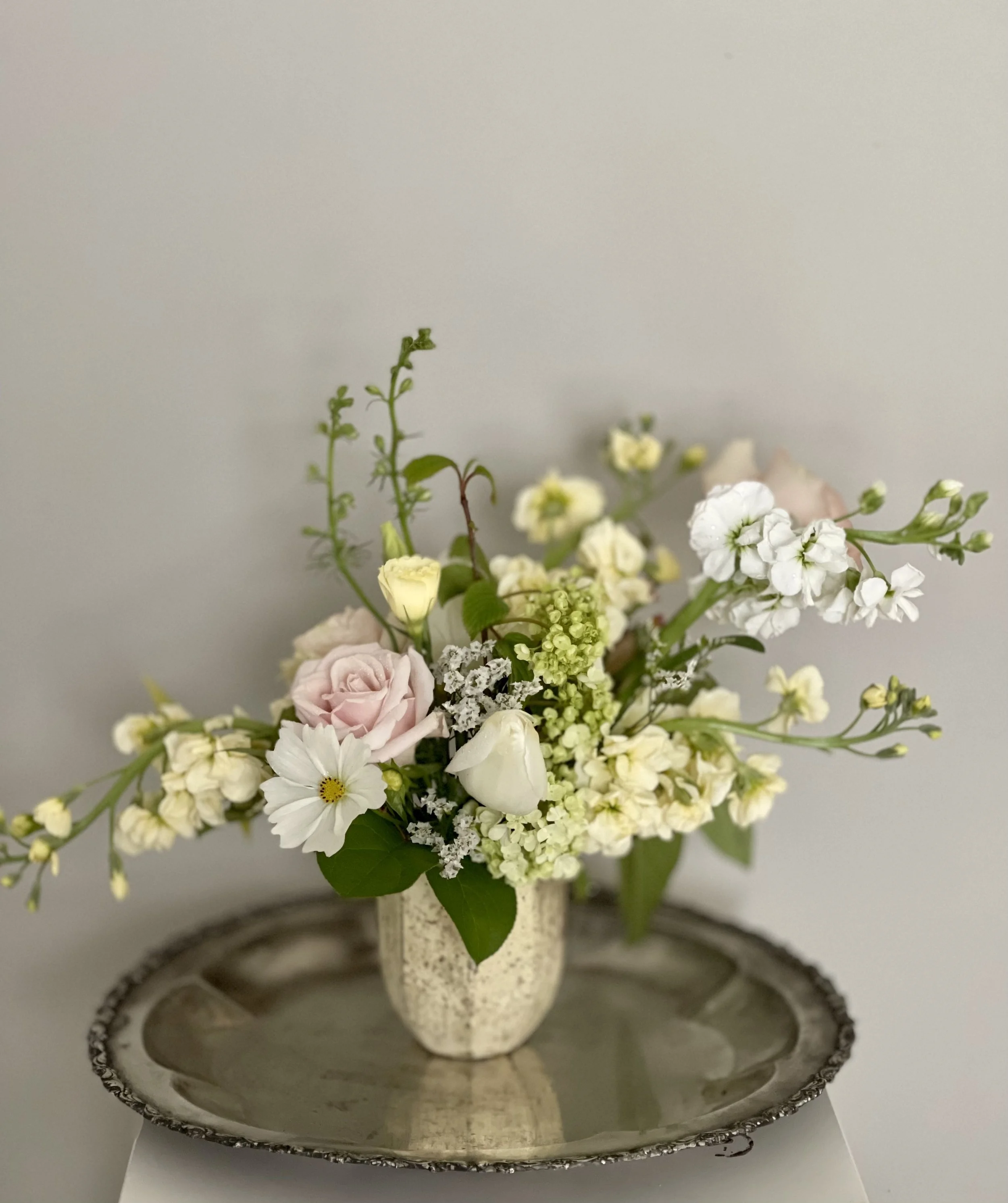 A floral arrangement with white, pink, and cream flowers in a small beige vase, placed on a silver tray against a light grey background.