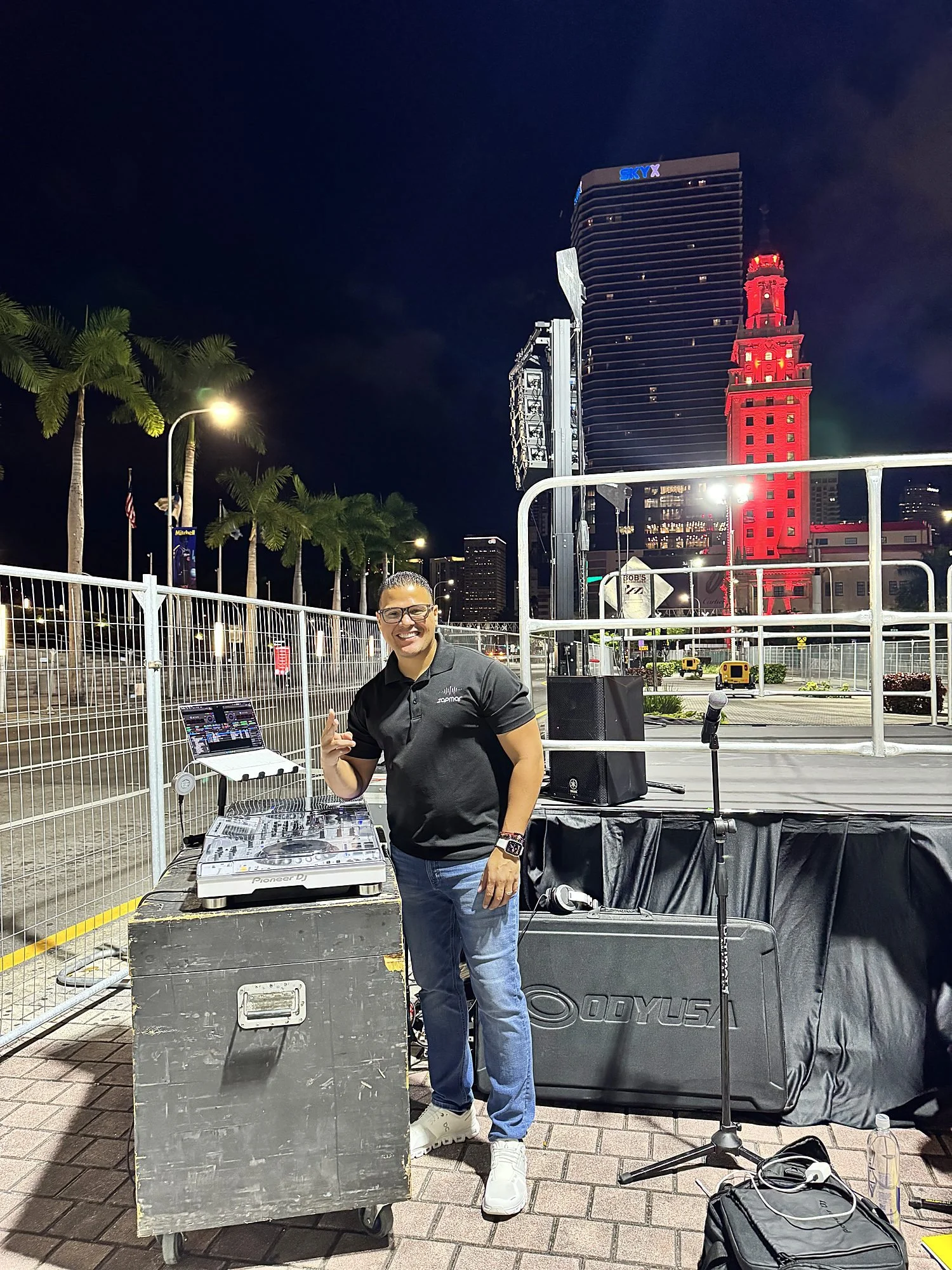 DJ Zapmar smiling beside his DJ booth at the Life Time Miami Marathon start line before sunrise, with the red lit Freedom Tower and downtown Miami skyline behind him