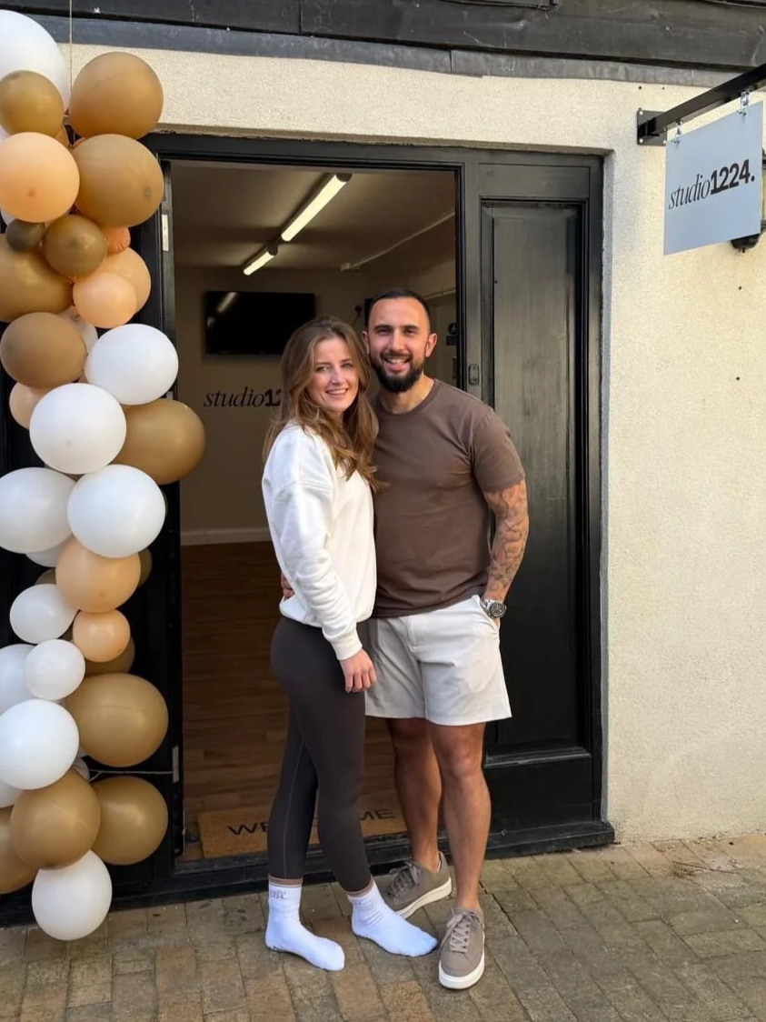 A smiling man and woman stand outside a studio entrance decorated with gold and white balloons. The woman wears a white hoodie and black leggings, and the man wears a brown t-shirt and white shorts.