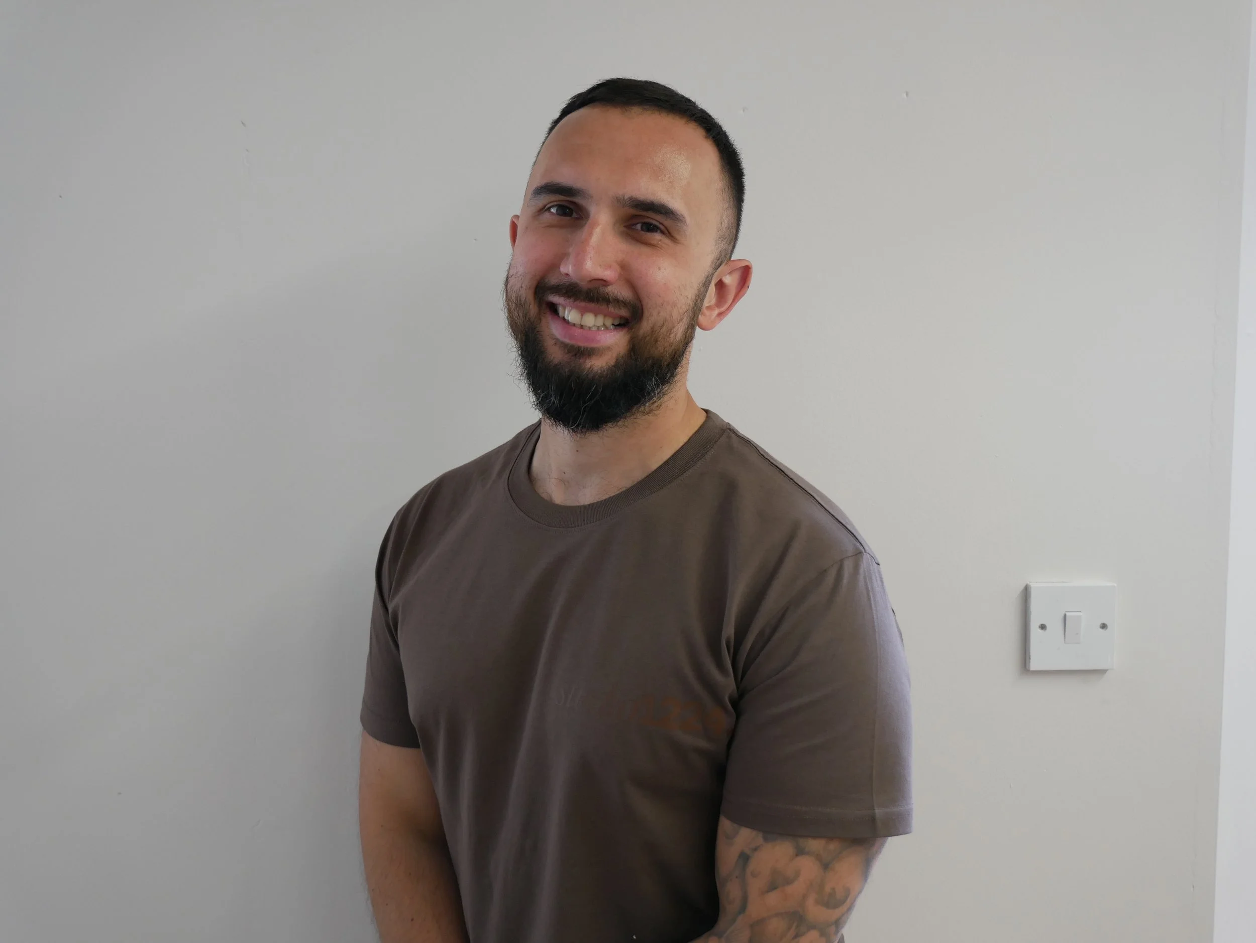 A man with a beard and short dark hair smiling and wearing a brown t-shirt, standing in front of a plain white wall.