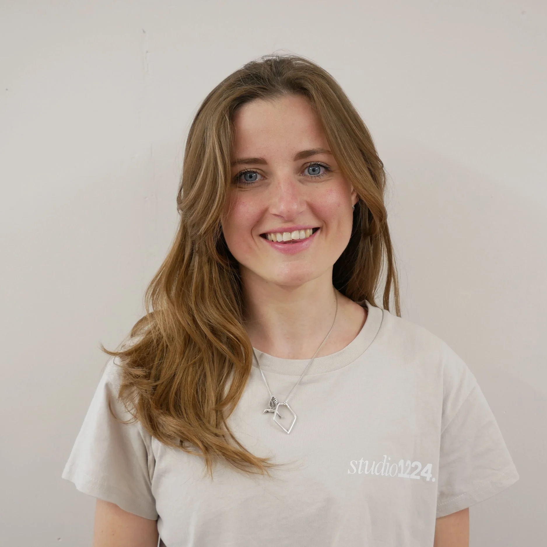A young woman with long, wavy, light brown hair, blue eyes, and fair skin smiling at the camera. She's wearing a beige T-shirt with white text that says "studio124" and a silver necklace with a geometric pendant. The background is a plain light-colored wall.